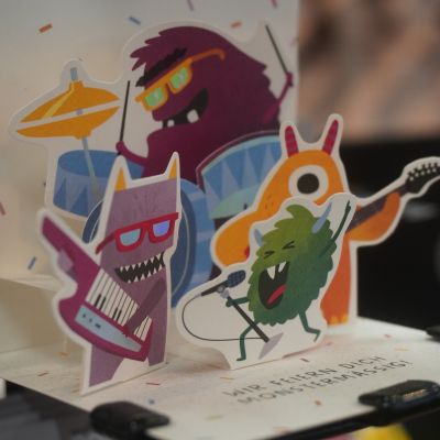 Eine Pop-up-Karte mit bunten Comic-Monstern, die als Rockband mit Schlagzeug, Gitarren und Mikrofon posieren. Darunter steht: „WIR FEIERN DICH MONSTERMÄSSIG!“
