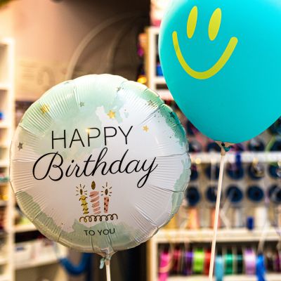 Zwei gefüllte Ballons am Stil. Links ein grün-weißer Ballon mit „HAPPY Birthday to you“ Aufschrift und rechts, etwas höher, ein türkiser Ballon mit gelbem Smiley
