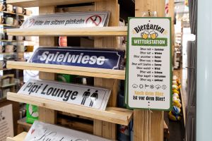 Verkaufsständer mit humorvollen Blechschildern für die Wand, darunter „Spielwiese“, „Pipi Lounge“ und eine „Biergarten Wetterstation“.