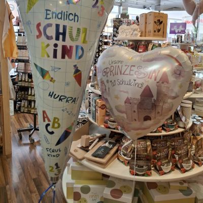 Zwei mit Gaß gefüllte Folienballons in Form einer Schultüte und eines Herzens, dahinter ein Etagentisch mit Geschenkartieln.
