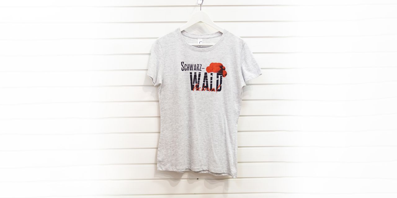 Weiß-graues T-Shirt mit Motiv, bestehend aus serifenlosem, texturiertem, schwarzem Aufdruck „Schwarz-WALD“, traditionellem roten Bollenhut und in roter Schreibschrift gesetzem „heimat“.