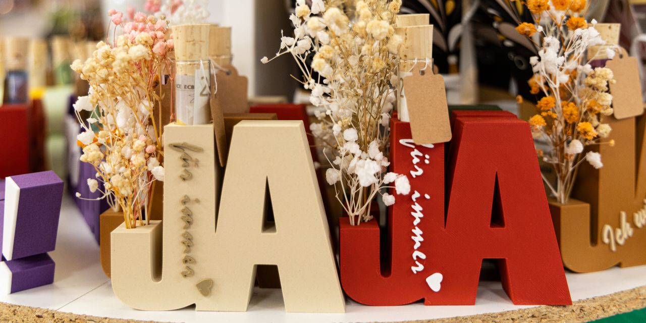 Dekorative Holzbuchstaben in Beige und Rot, die das Wort „JA“ formen, verziert mit Trockenblumen und dem Schriftzug „für immer“.