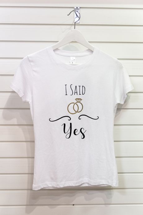 Weißes T-Shirt mit schwarzem Aufdruck "I SAID Yes", umgeben von schwarzen Ornamenten und stilisierten goldenen Ringen, die wie Kettenglieder miteinander verbunden sind. Einer der Ringe trägt einen Diamanten.