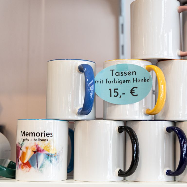 Stapel weißer Keramiktassen mit verschiedenfarbigen Henkeln im Regalfach. Ein Preisschild zeigt „Tassen mit farbigem Henkel 15,- €“.