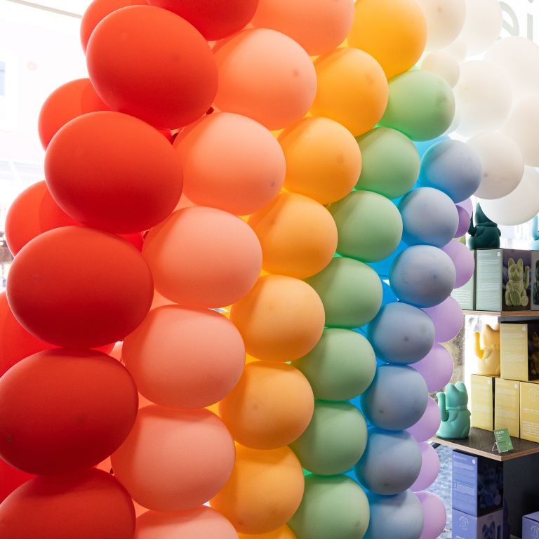 Farbenfrohe Luftballongirlande in Regenbogenfarben von Rot über Gelb und Grün bis Blau in einem Verkaufsraum.