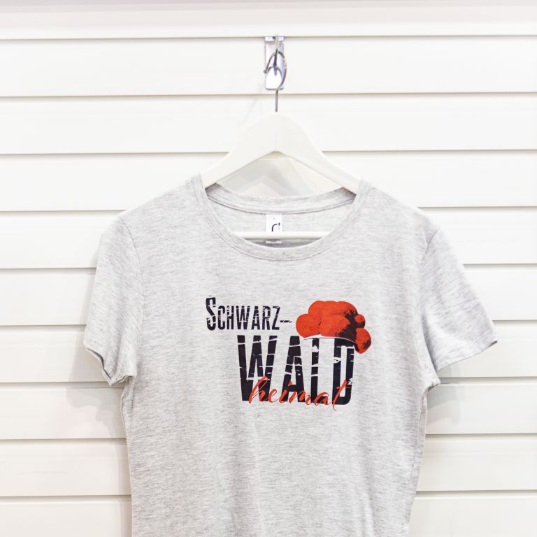 Weiß-graues T-Shirt mit Motiv, bestehend aus serifenlosem, texturiertem, schwarzem Aufdruck „Schwarz-WALD“, traditionellem roten Bollenhut und in roter Schreibschrift gesetztem „heimat“.