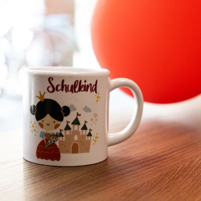 Eine Keramiktasse mit der Aufschrift „Schulkind“ und einer Grafik, die ein kleines Mädchen mit Schultüte vor einem Gebäude mit vielen Türmen zeigt.
