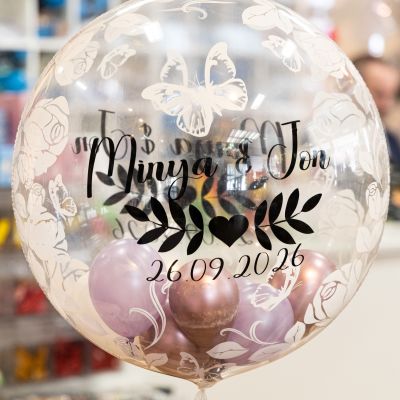 Ein durchsichtiger, personalisierter Hochzeitsballon mit der Aufschrift „Minya & Jon 26.09.2026“, gefüllt mit kleinen lila-goldenen Ballons.