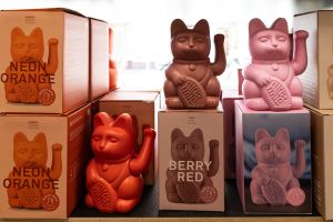 Winkekatzen in verschiedenen Neon- und Mattfarben wie Orange, Beere und Rosa neben den dazugehörigen Produktverpackungen.