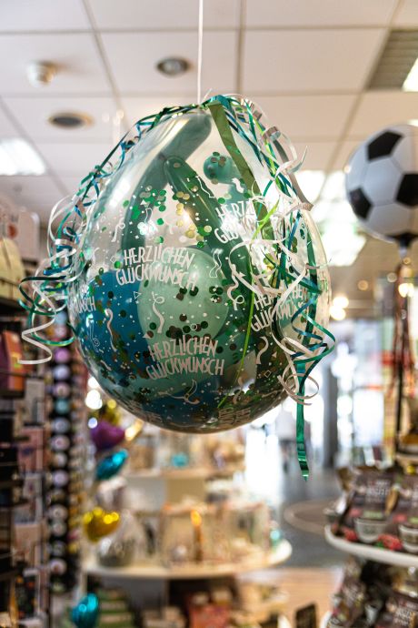Mit grünenn Bändern dekorierter, gefüllter transparenter Ballon mit Inhalt. Bedruckt „Herzlichen Glückwund“.